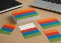 Colorful printed cards arranged on a wooden table with a laptop and notebook in the background.
